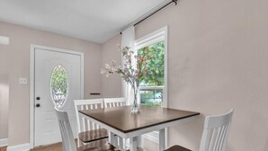 Ein Esszimmer mit Holztisch und weißen Stühlen, einer Vase mit Blumen, einer Tür mit bleiverglastem Fenster und Blick ins Grüne.
