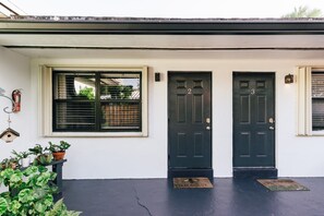 A house with two black doors, a window with blinds, and a small porch area with potted plants.