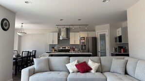 Un salon moderne avec un canapé gris, un coin repas avec une table et des chaises, et une cuisine équipée d’appareils électroménagers en acier inoxydable.