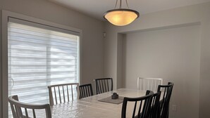 Un coin repas avec une table recouverte de plastique, six chaises, un luminaire suspendu et une fenêtre avec des stores.