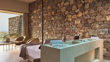 A modern bedroom with a stone wall, a large bed, a built-in sink, and a view of the outdoors.