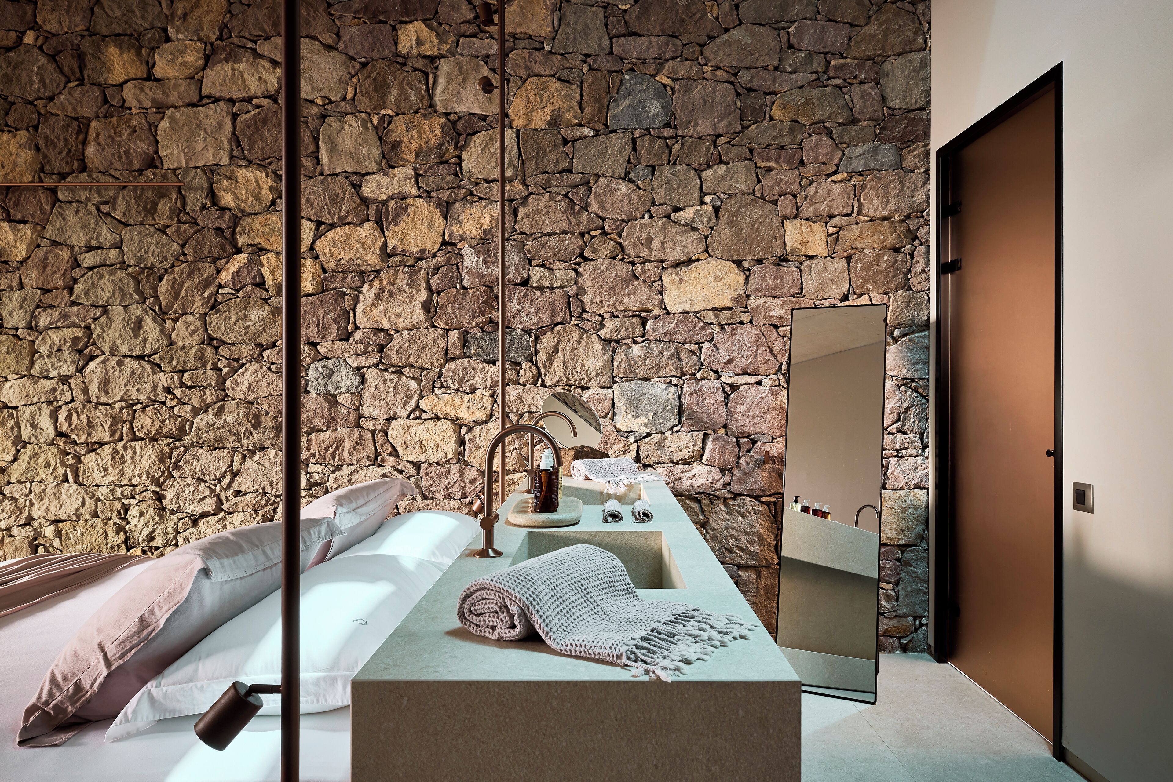 Un baño moderno con pared de piedra, un espejo grande, un lavabo y una cama con toalla y almohada.