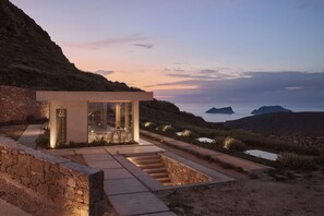 Um edifício moderno com fachada de vidro, localizado numa encosta com vista para o mar e ilhas.