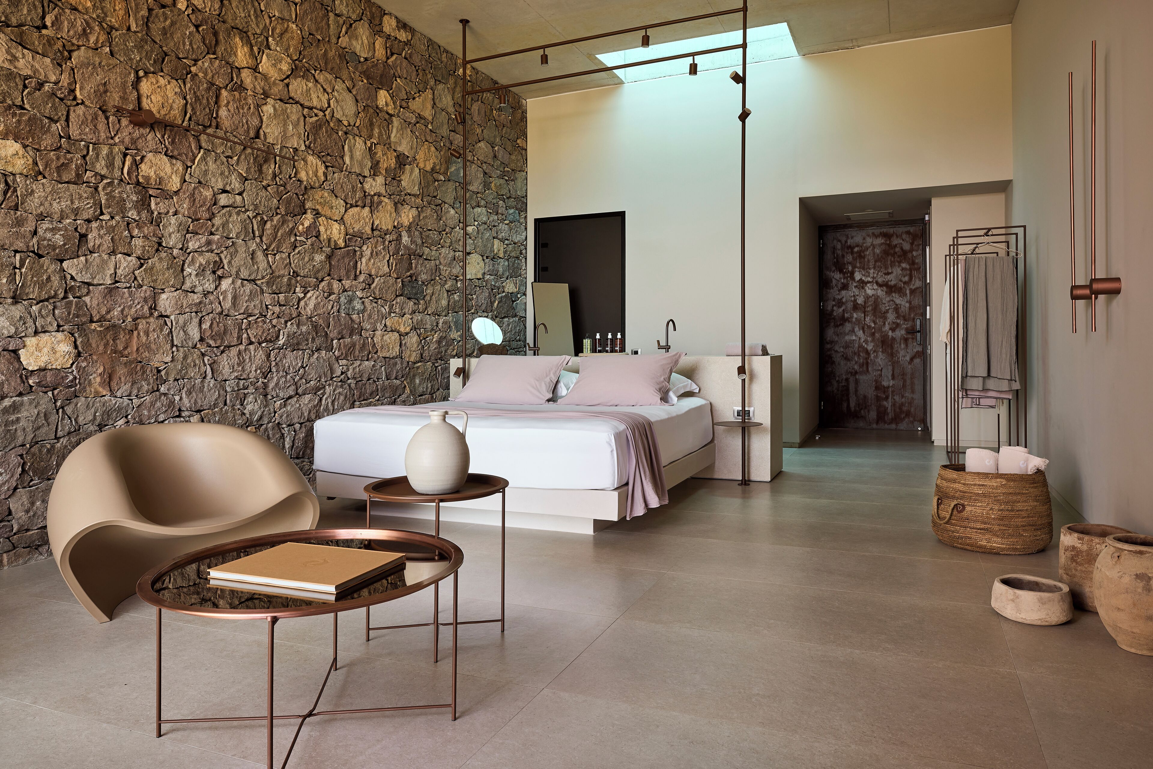 Un dormitorio moderno con pared de piedra, una cama con ropa blanca, una silla, una mesita de noche y una lámpara colgante.