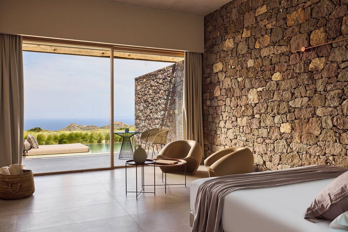A bedroom with a stone wall, a bed, a table with chairs, and a view of the outdoors.