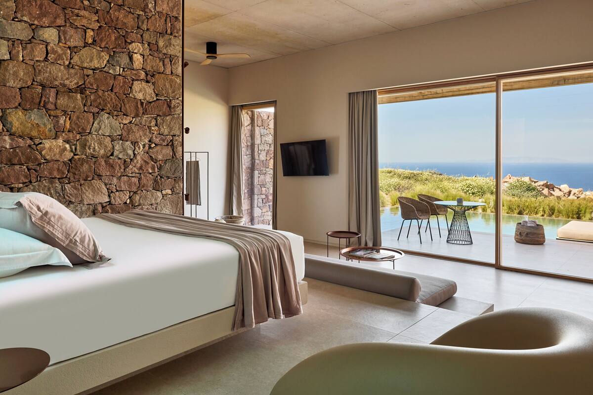 A bedroom with a stone wall, a bed, a TV, and a view of the ocean.
