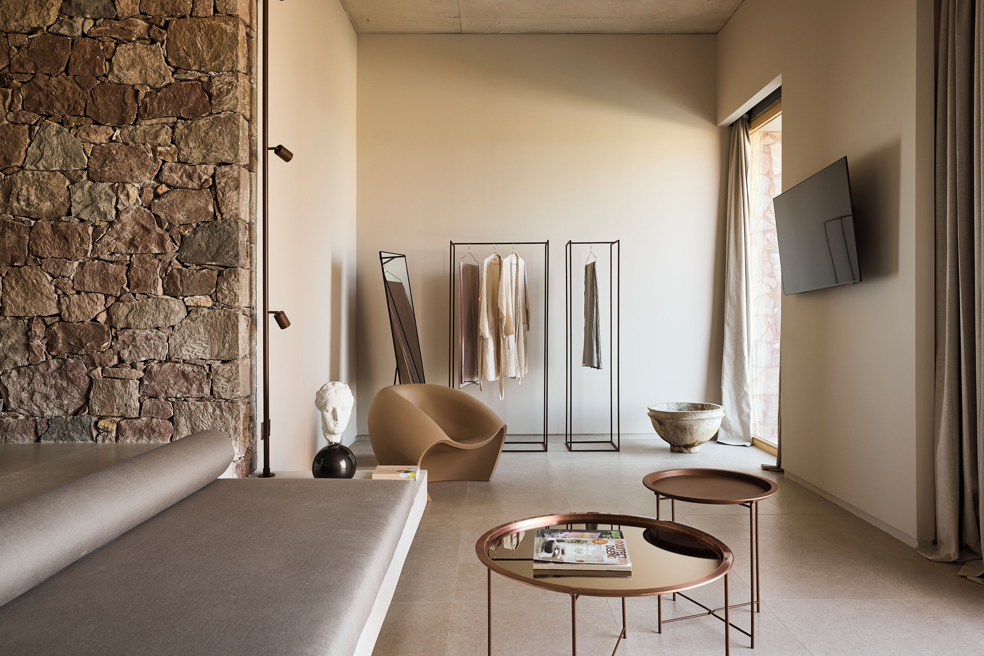 A modern bedroom with a stone wall, a bed, a curved chair, a TV, and a window with curtains.