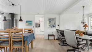 Uma ampla área de refeições com cadeiras de madeira e uma mesa, uma cozinha com armários brancos e vista para o exterior.