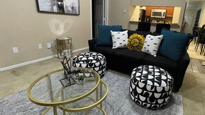 Una sala de estar moderna con un sofá negro, otomanas estampadas, una mesa de centro de vidrio y un cuadro en la pared.