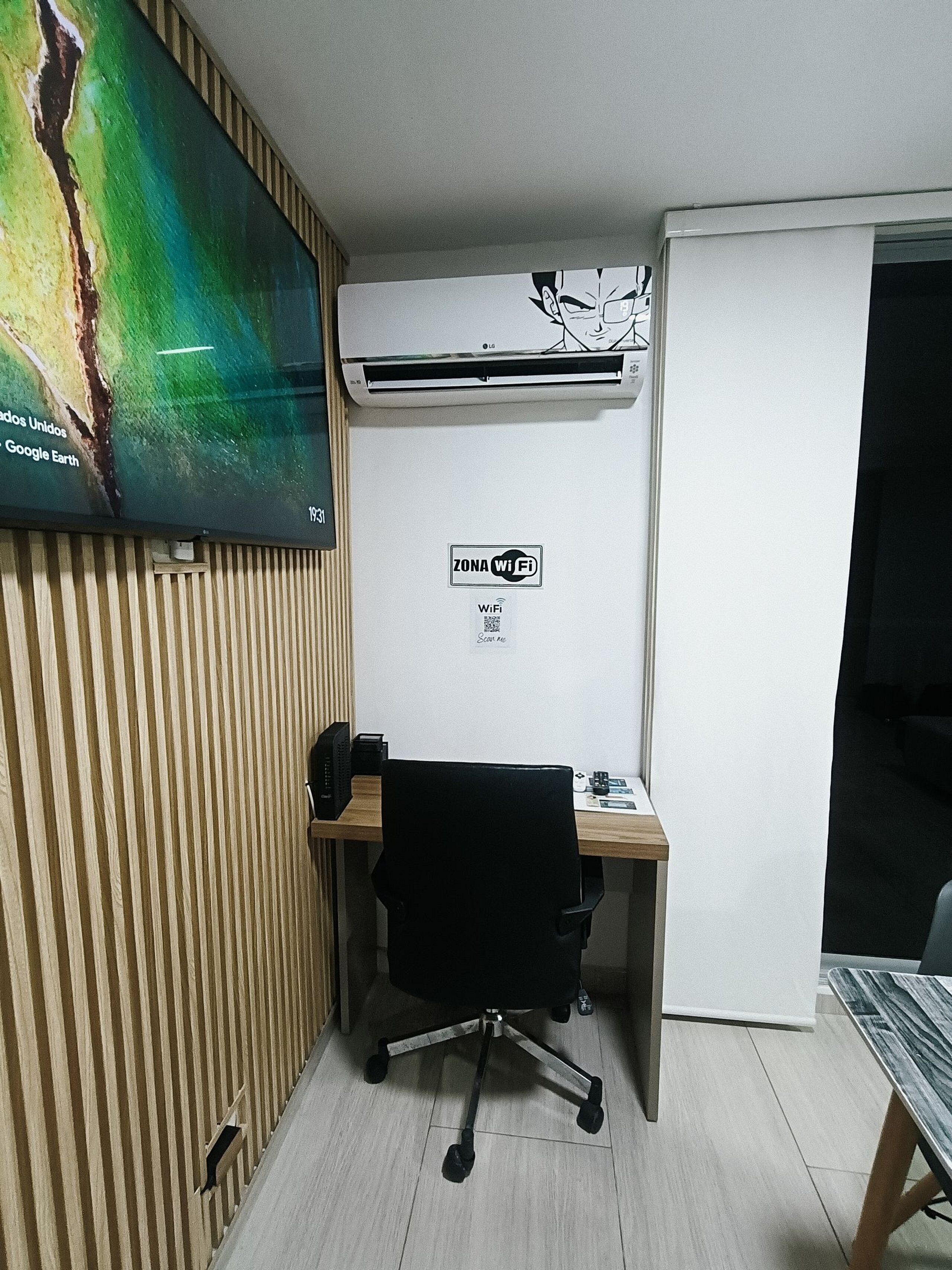 Un piccolo ufficio con parete in listelli di legno, condizionatore fissato al muro, una scrivania con sedia e una TV che mostra una mappa.