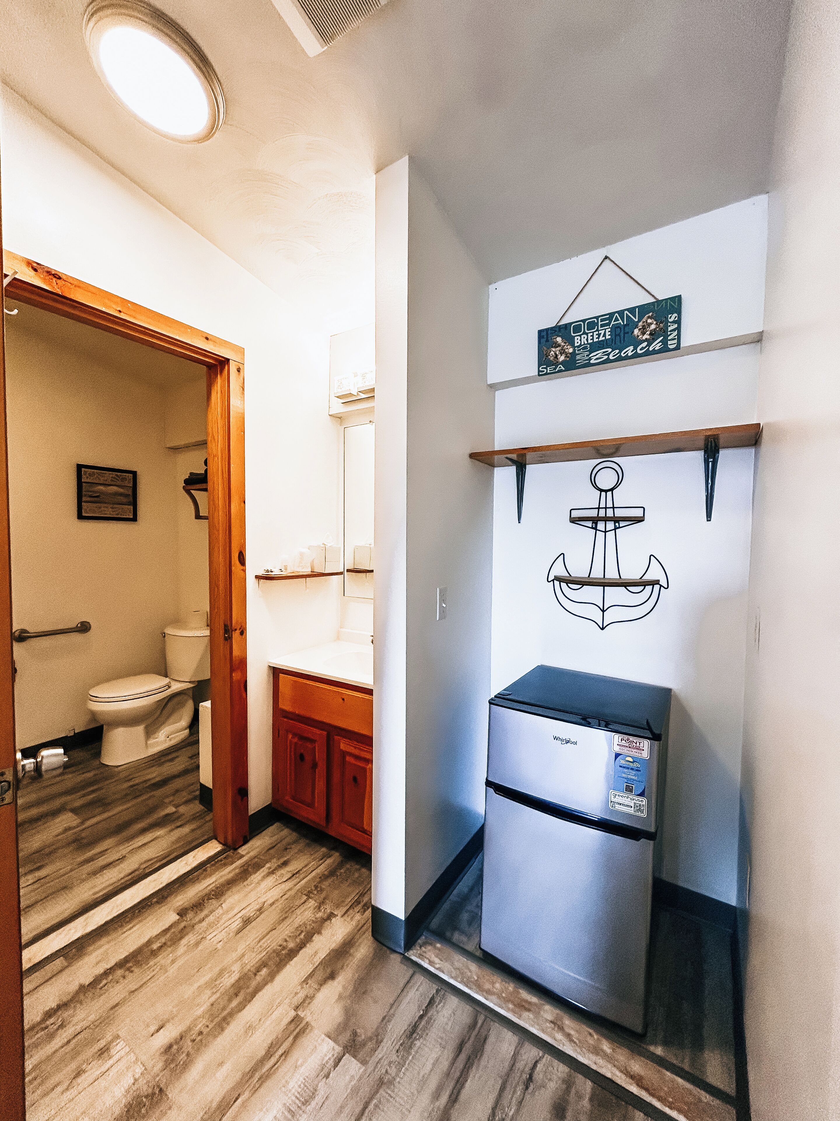 Un baño con inodoro, armarios de madera, un refrigerador de acero inoxidable y un estante en la pared con un letrero de temática náutica.