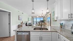 A modern kitchen with white cabinets, a dishwasher, and a large island with a sink. There is a dining area with a table and chairs, and a view of the ocean through large windows. - Romar Place 1106 (Orange Beach)