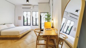Une chambre avec un lit, un bureau en bois et une chaise. Il y a une fenêtre avec des rideaux, une plante et un miroir accroché au mur.