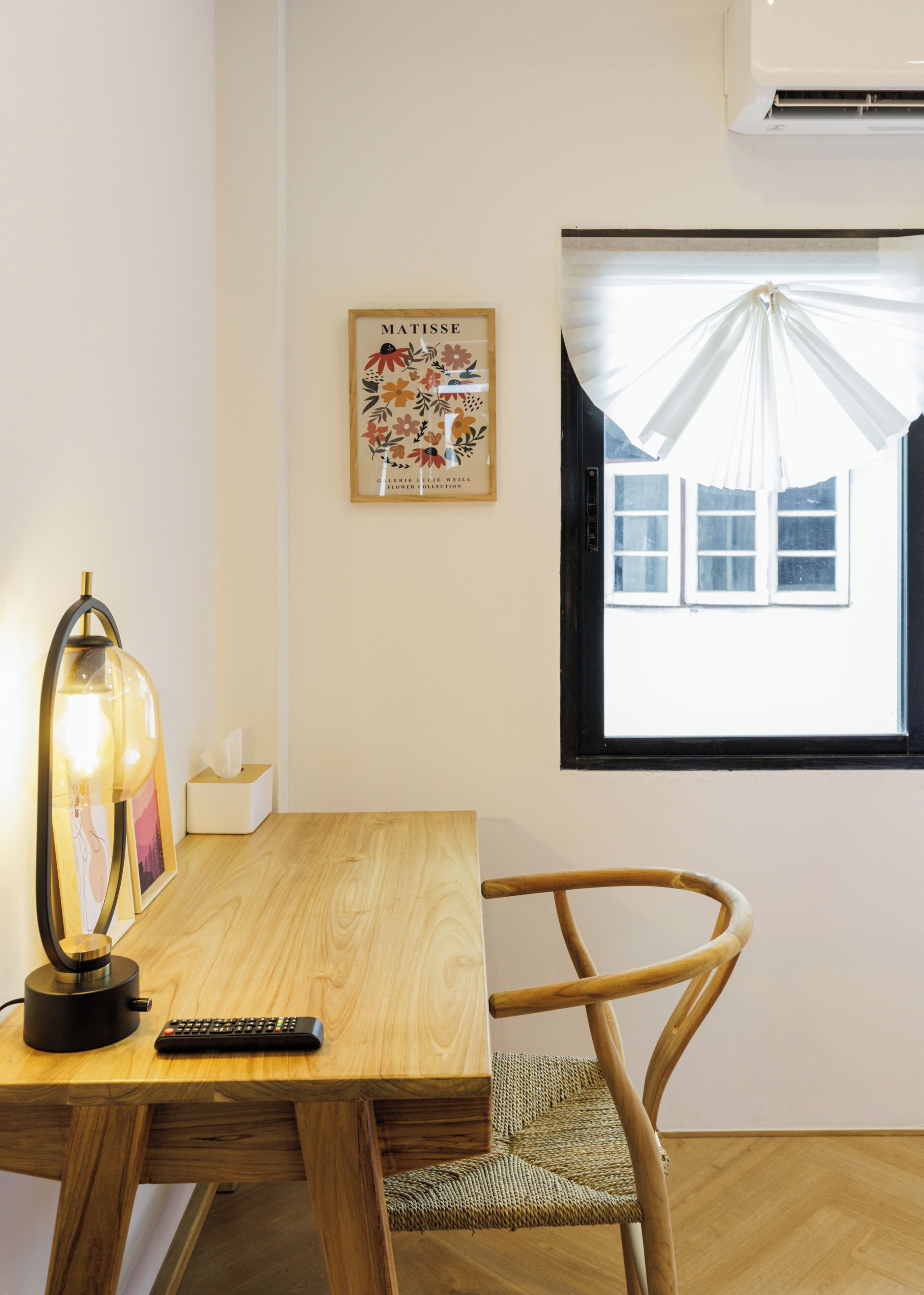 Ett rum med ett skrivbord i trä, en stol, en lampa, en fjärrkontroll, ett inramat konstverk och ett fönster med en gardin.