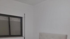 Ein ordentlich gemachtes Bett mit weißen Leinen und Kissen, ein Fenster mit Jalousien und ein Holzboden.