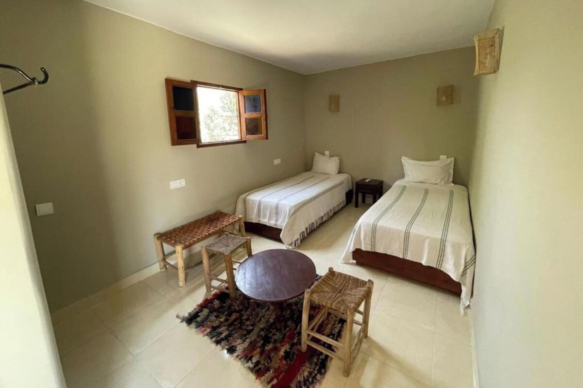 A bedroom with two single beds, a small table, a bench, and a window.