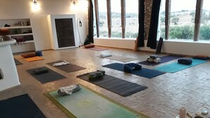 A yoga studio with mats and cushions arranged on a wooden floor, large windows with curtains, and a shelf with books and a water bottle. - Tasguayan (Tafedna)