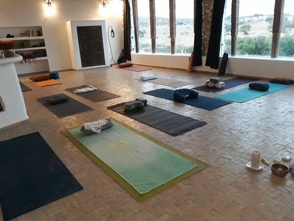 Un studio de yoga avec des tapis et des coussins disposés sur un plancher en bois, de grandes fenêtres avec des rideaux et une étagère contenant des livres et une bouteille d’eau.