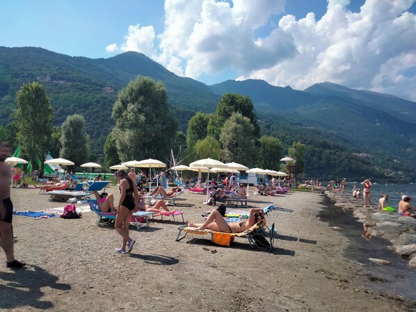 A beach with sun loungers and umbrellas, surrounded by mountains and trees.