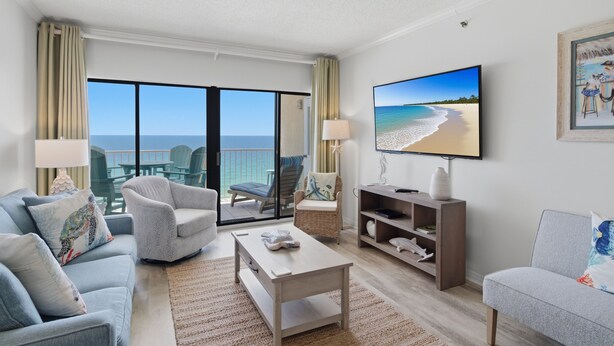 A living room with a large TV, a sofa, a coffee table, and a view of the beach. - September Dates Available! Tropical Winds 703! (Gulf Shores)