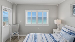 A bedroom with a bed, a nightstand with a lamp, a window with shutters, and a chair. - September Dates Available! Tropical Winds 703! (Gulf Shores)