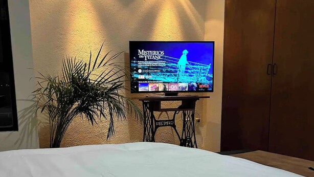 A bed with a remote control in the foreground, a television displaying 'Misterios del Titanic' on a wall-mounted stand, and a plant to the left.