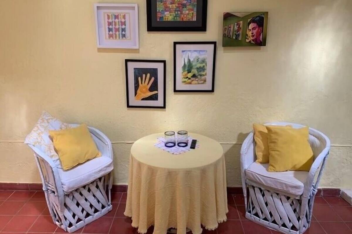 A small room with two white wicker chairs with yellow cushions, a round table with a white tablecloth, and four framed artworks on the wall.