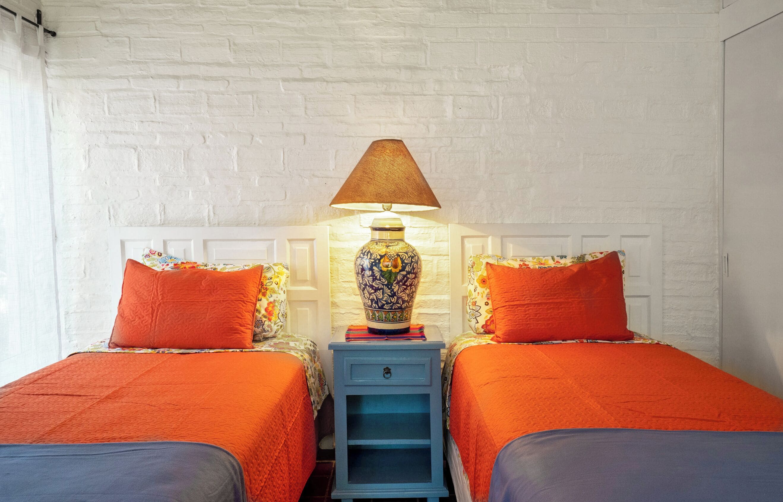 A room with two beds, each with an orange bedspread, a matching orange pillow, and a decorative vase on a nightstand between them.