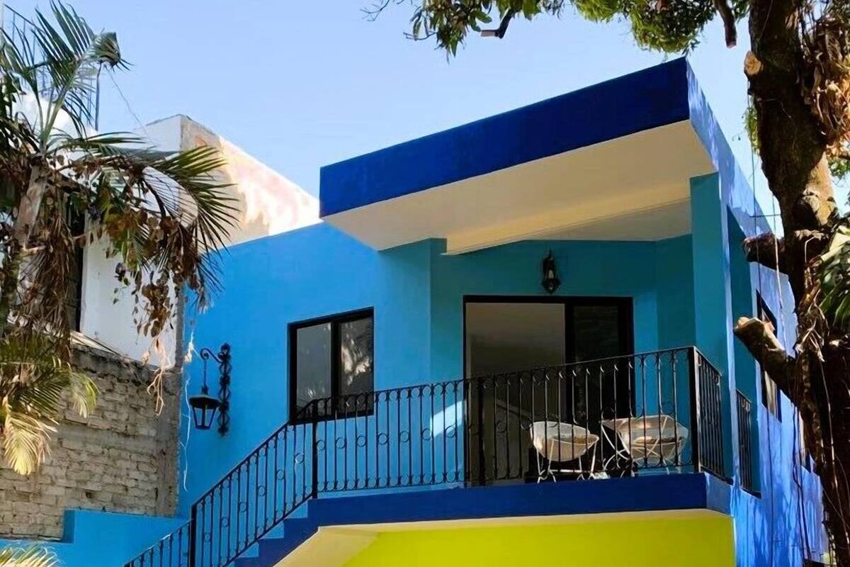 A two-story building with a blue and yellow facade, a balcony with a metal railing, and a staircase leading to the upper floor.