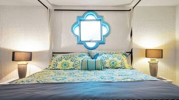 Una cama con colcha estampada, dos lámparas de noche y un espejo con marco decorativo fijado a la pared.