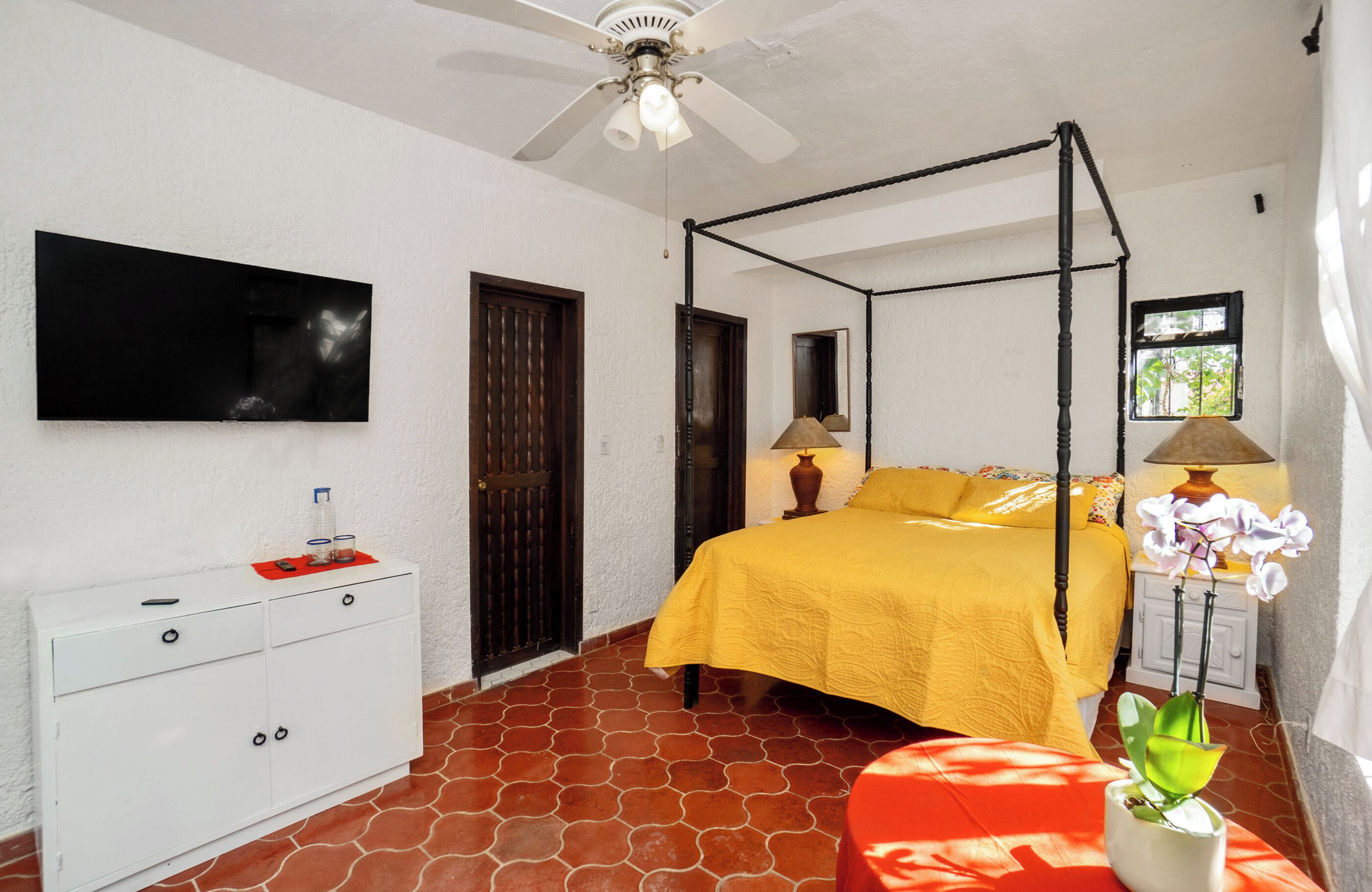 Un dormitorio con cama con dosel, ventilador de techo, televisor montado en la pared, cómoda blanca y ventana con vistas a árboles.