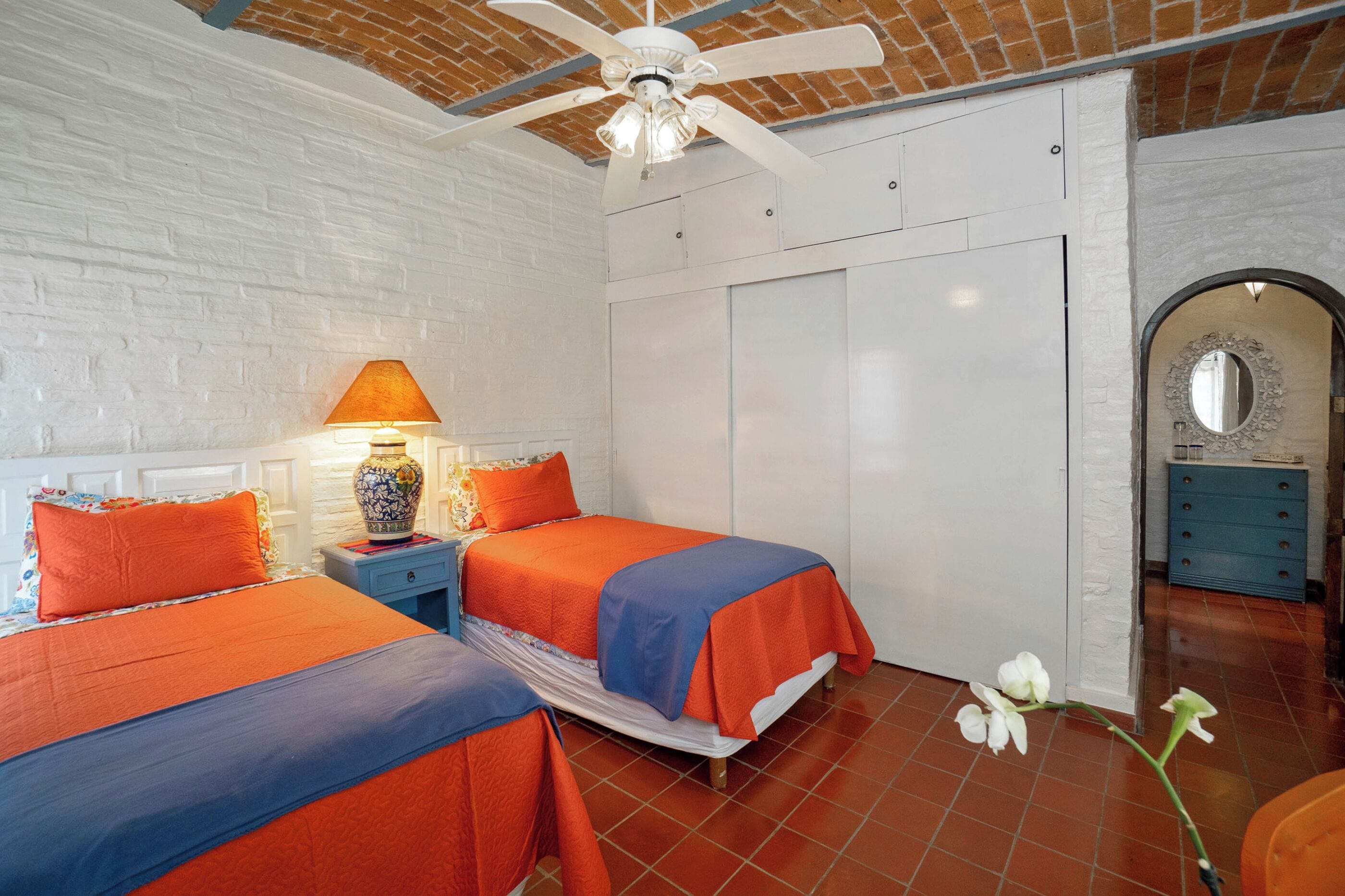 Un dormitorio con dos camas, un ventilador de techo, una mesita de noche con lámpara, un armario y un espejo decorativo.