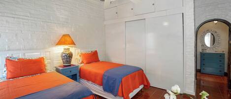 Un dormitorio con dos camas, un ventilador de techo, una mesita de noche con lámpara, un armario y un espejo decorativo.