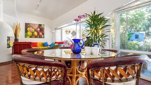 A dining area with a glass table, wicker chairs, a colorful painting, and a large window with a view of greenery. - Garden of Dreams Boutique Hotel (Chapala)