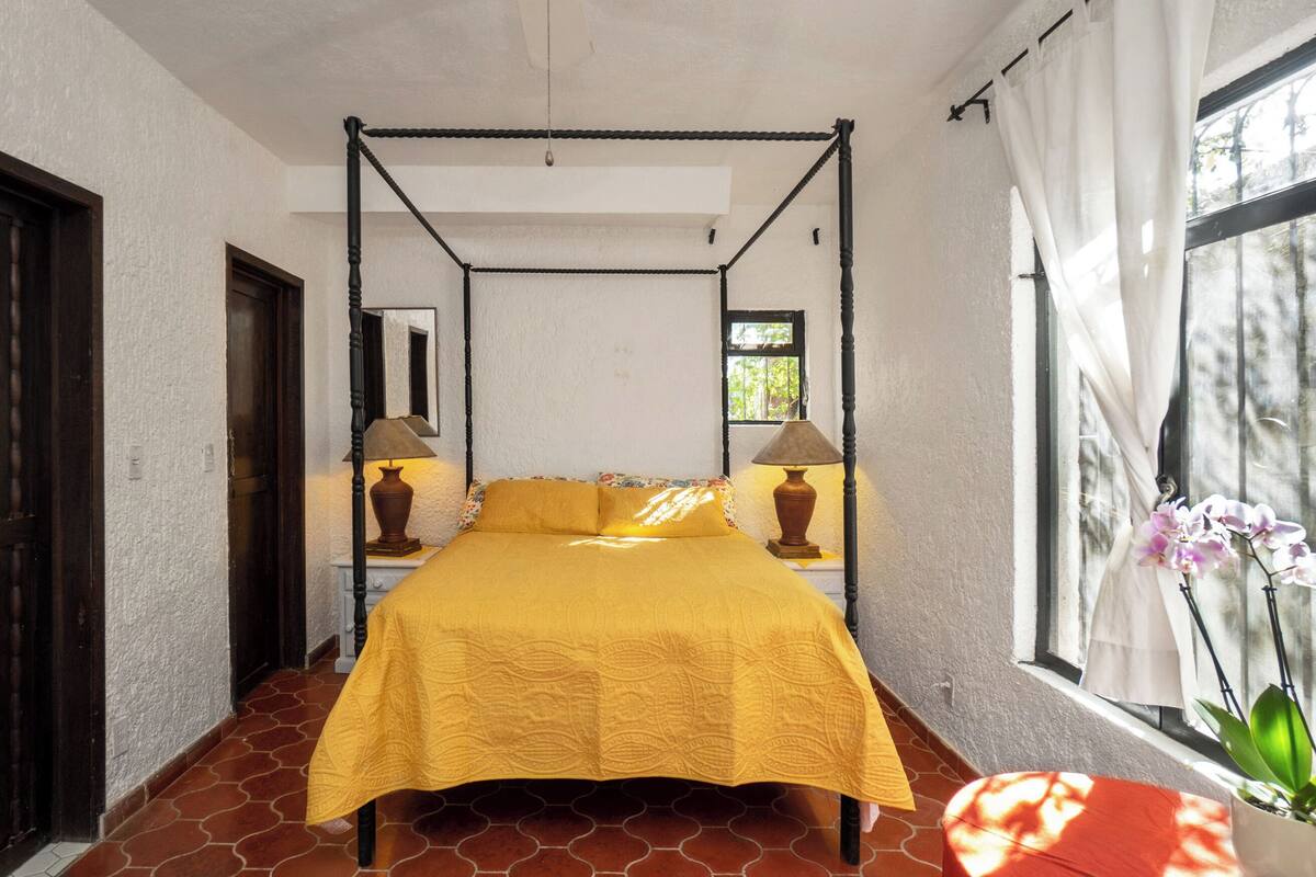 A room with a four-poster bed, two bedside lamps, a window with curtains, and a potted plant.