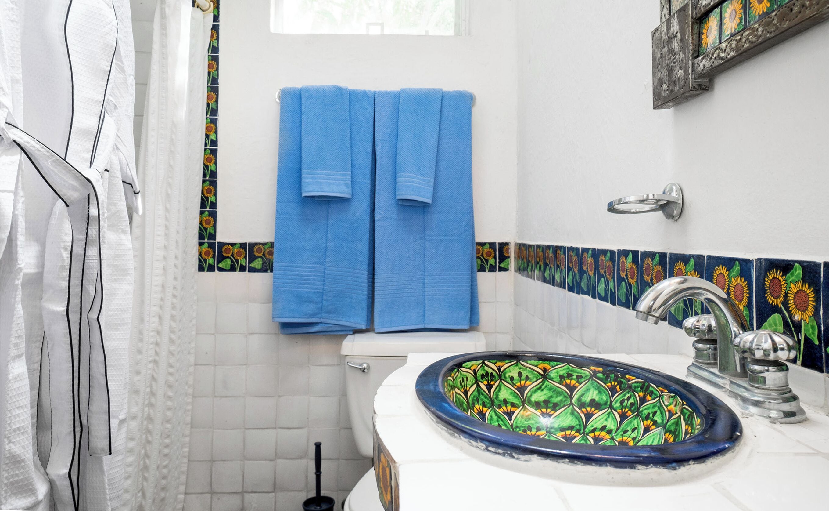 Une salle de bain avec des toilettes blanches, un porte-serviettes bleu avec deux serviettes, un lavabo avec un receveur décoratif en carrelage et un rideau de douche orné d’un motif de tournesols.