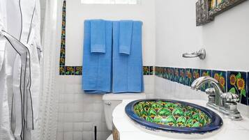 Une salle de bain avec des toilettes blanches, un porte-serviettes bleu avec deux serviettes, un lavabo avec un receveur décoratif en carrelage et un rideau de douche orné d’un motif de tournesols.