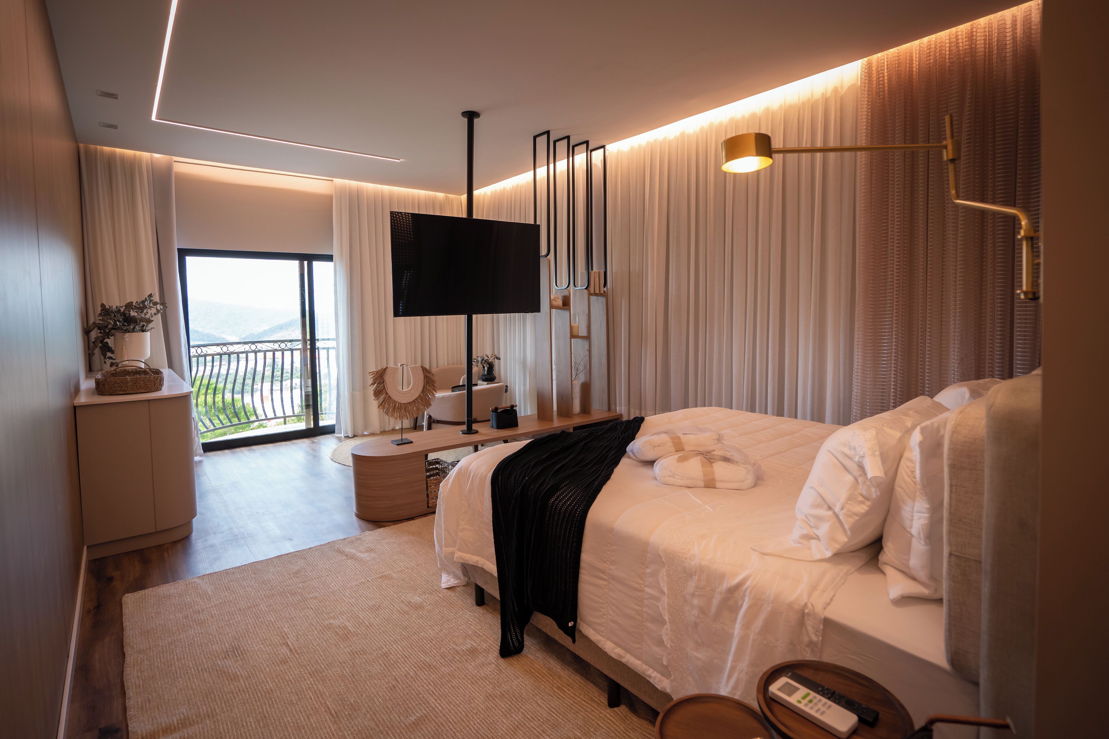 Quarto de hotel moderno com uma cama grande, televisão, mesa de cabeceira, abajur e vista para o exterior.