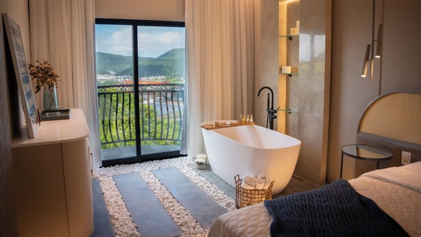 A modern bathroom with a freestanding bathtub, a small bedside table, and a view of a balcony with a railing. - Morada Pedra Branca  (Balneário Camboriú)