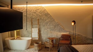 A modern bathroom with a freestanding bathtub, a wooden floor, a stone wall, a towel rack, a wicker basket, and a floor lamp. - Morada Pedra Branca  (Balneário Camboriú)