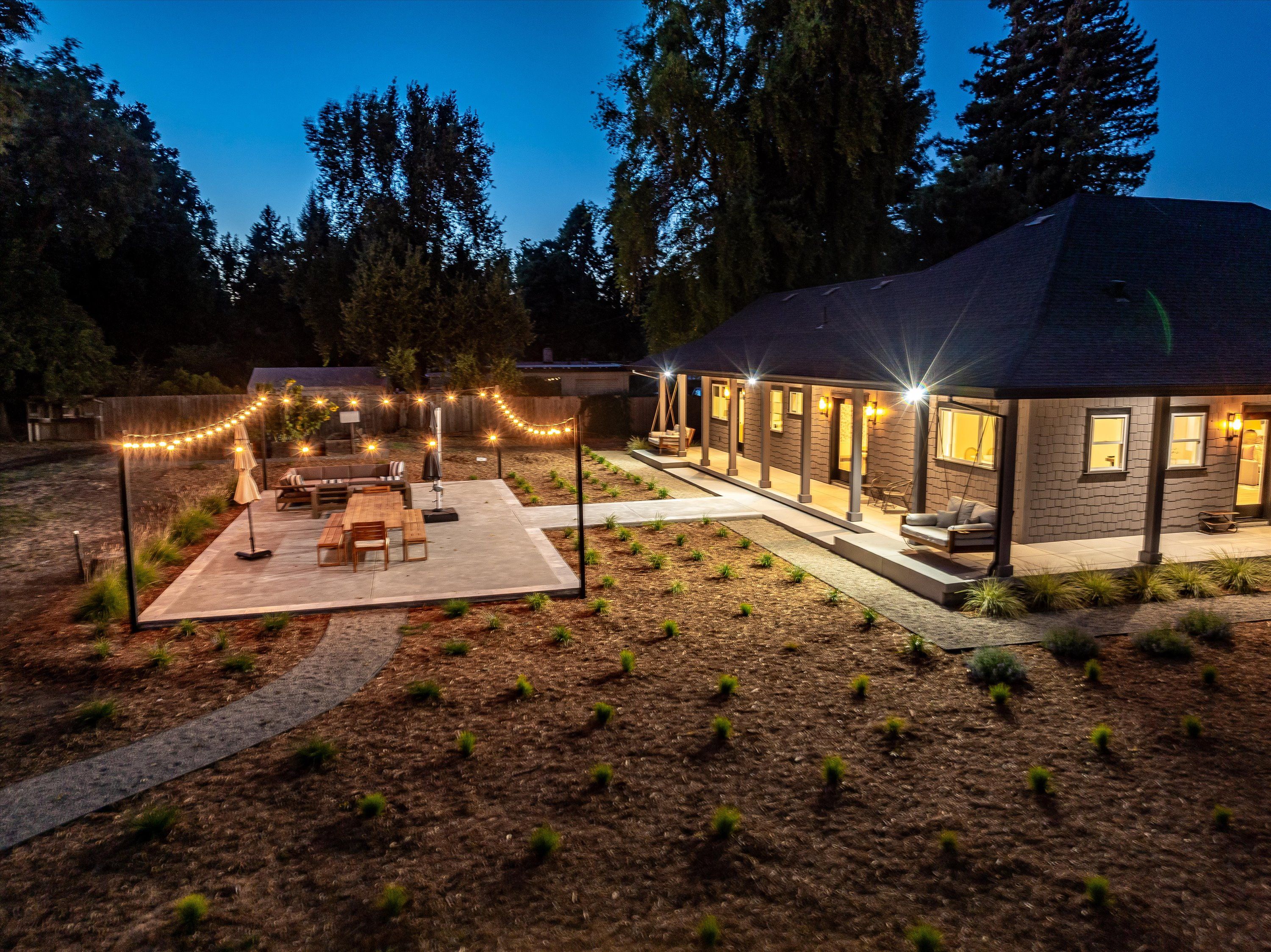 Een overdekt terras buiten met eethoek, lichtslingers, en een huis met een stenen gevel en grote ramen.