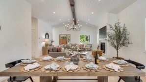 A spacious dining area with a wooden table set for six, surrounded by modern furniture and a large vase of flowers.