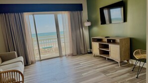 Una habitación con piso de madera, un sofá, una mesa de comedor, un televisor en la pared y vista a la playa a través de puertas correderas.