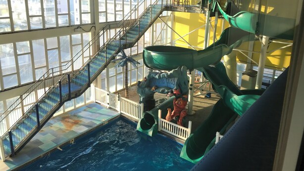 Un parque acuático interior con una piscina grande, varias toboganes y una escalera que lleva a niveles superiores.