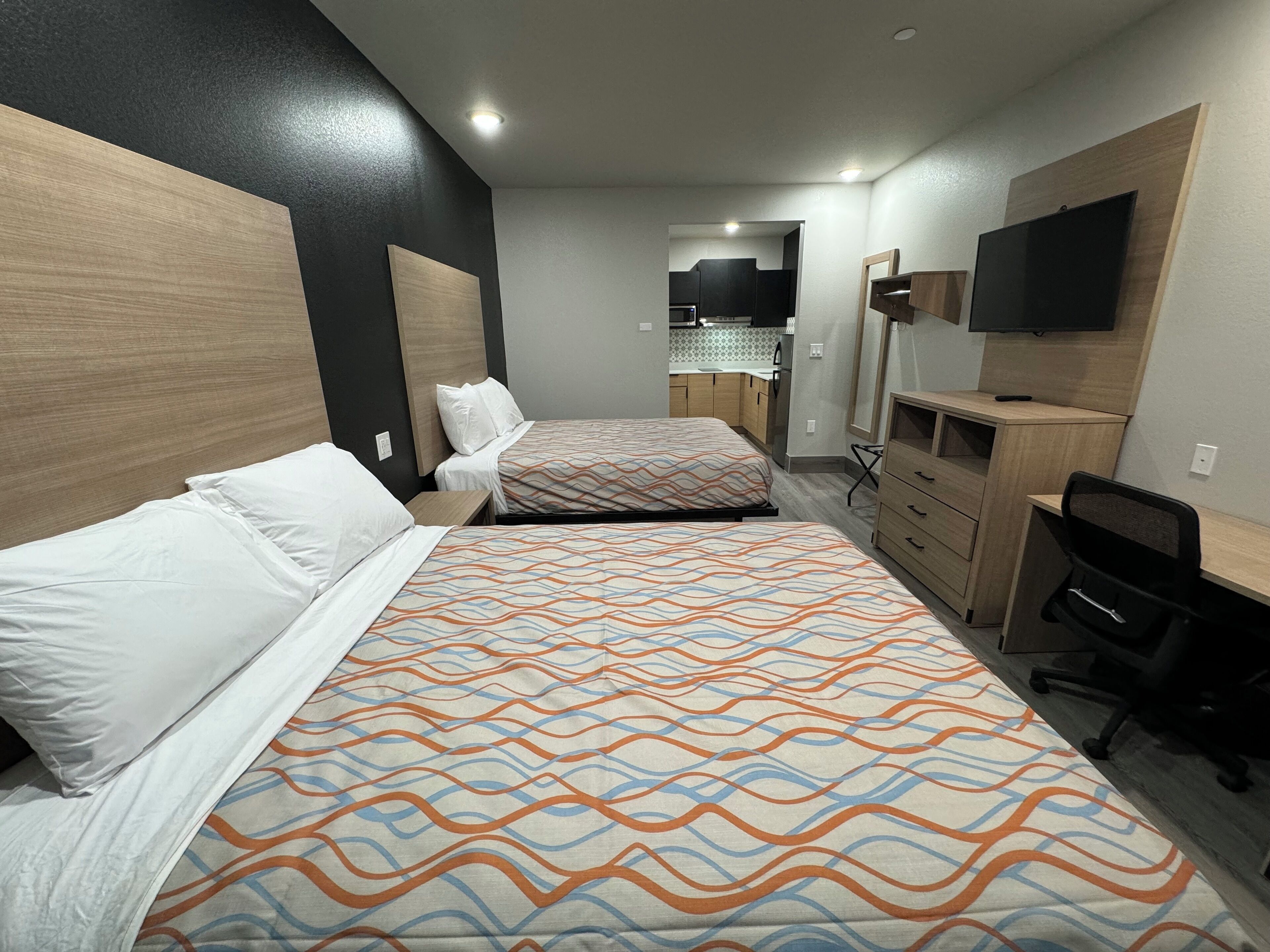 A hotel room with two beds, a desk, a chair, a TV, and a kitchenette in the background.