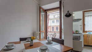 Een eetgedeelte met een houten tafel gedekt voor twee personen, een vaas met gele bloemen, en uitzicht op de keuken en het balkon.