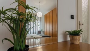 Een moderne kamer met een houten vloer, een grote spiegel die een bed en een raam reflecteert, en twee kamerplanten.