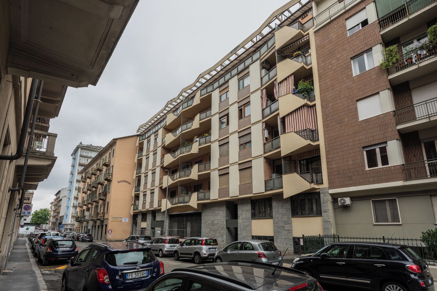 En flereetagers boligbygning med altaner, en parkeringsplads med biler og et gadebillede.