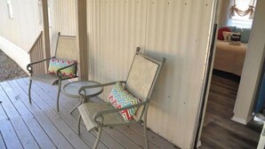 En veranda med to metalstole og et lille bord, en trædør, og et soveværelse, der kan ses igennem døren.