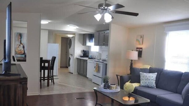 Sala de estar com sofá, mesa de centro e televisão em móvel. Área de jantar com cadeiras e mesa. Cozinha visível ao fundo, com armários e eletrodomésticos.